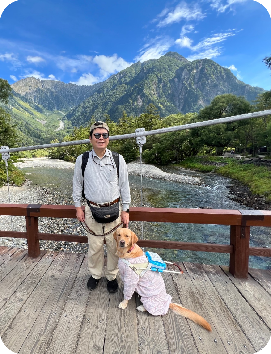 青空と白い雲の下、山と川を背景にして橋の上で笑顔の松井さんとウパちゃん