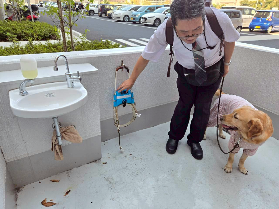 外の水道の横で壁にかけている盲導犬のリードに触れる松井さん